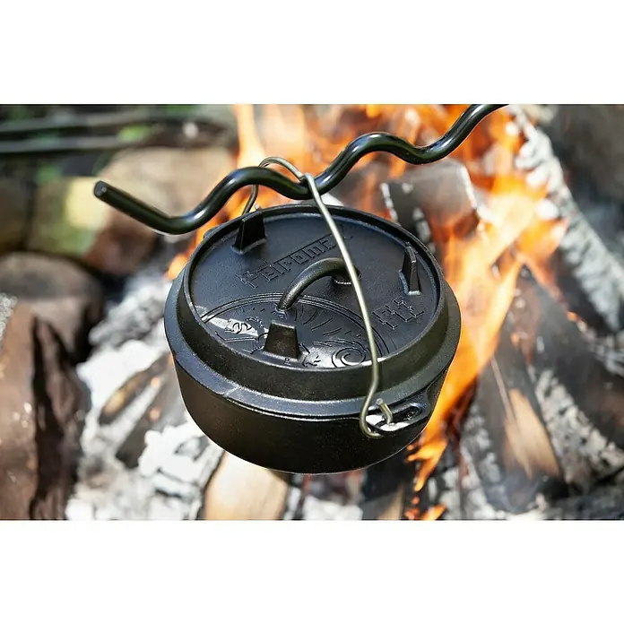 Petromax Dutch Oven - Ø x H: 25,2 x 12 cm, Gusseisen, 1,6 l, Planer Boden - Ein gusseiserner Dutch Oven hängt über einem Lagerfeuer, ideal zum Kochen im Freien. Das robuste Kochgeschirr ist perfekt für Camping und Outdoor-Abenteuer.