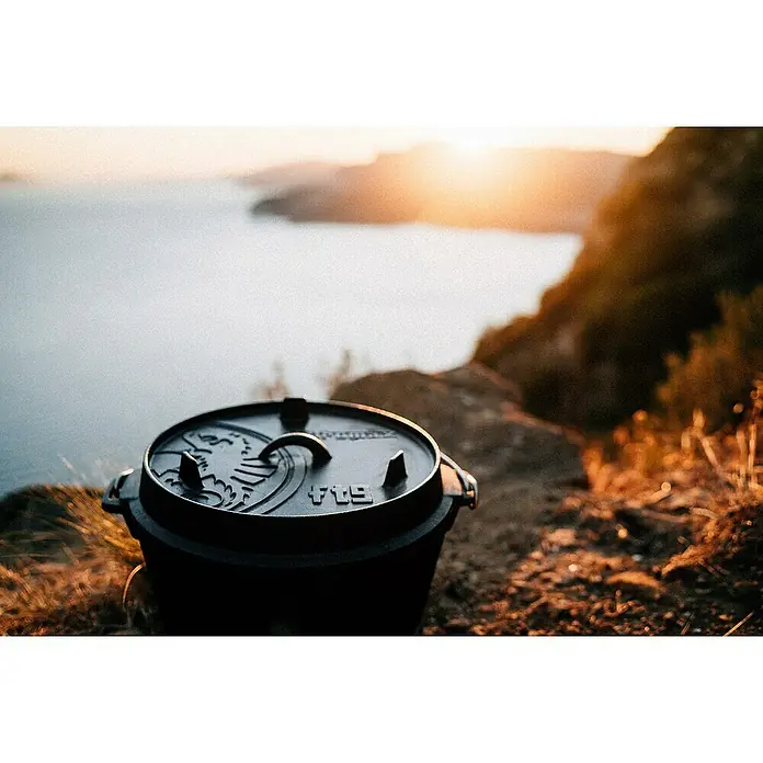 Petromax Dutch Oven - Ø x H: 36,4 x 19,2 cm, Gusseisen, 7,5 l, Planer Boden - Ein schwarzer Dutch Oven mit Deckel steht auf einem Felsen mit Blick auf das Meer bei Sonnenuntergang.