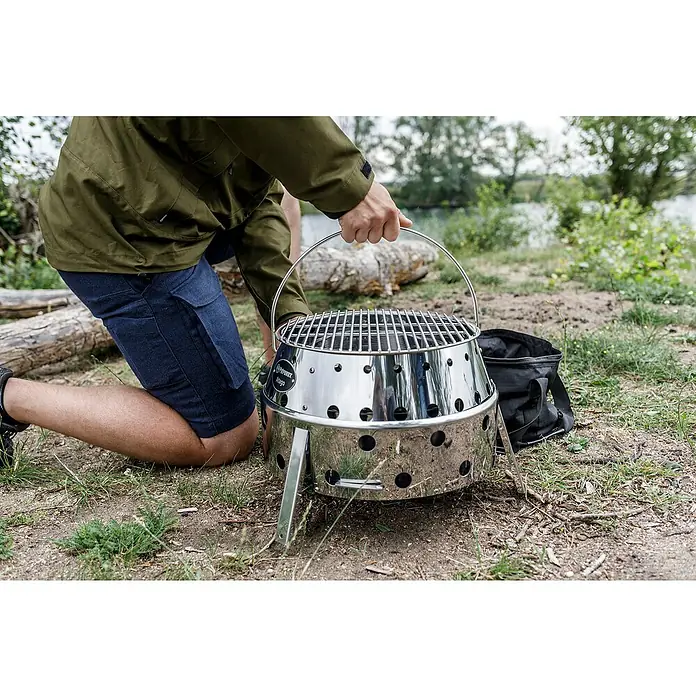 Petromax Holzkohlegrill Atago - Grillfläche (B x T): 45,1 x 44,5 cm, Durchmesser Grillfläche: 34 cm, Silber - Person hockt neben einem Atago Grill von Petromax. Der Grill ist tragbar, aus Edelstahl und hat eine runde Form mit Löchern.