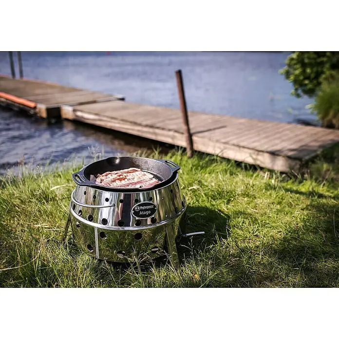 Petromax Holzkohlegrill Atago - Grillfläche (B x T): 45,1 x 44,5 cm, Durchmesser Grillfläche: 34 cm, Silber - Ein Petromax Atago Grill mit Speck in einer Pfanne steht am Ufer eines Sees.