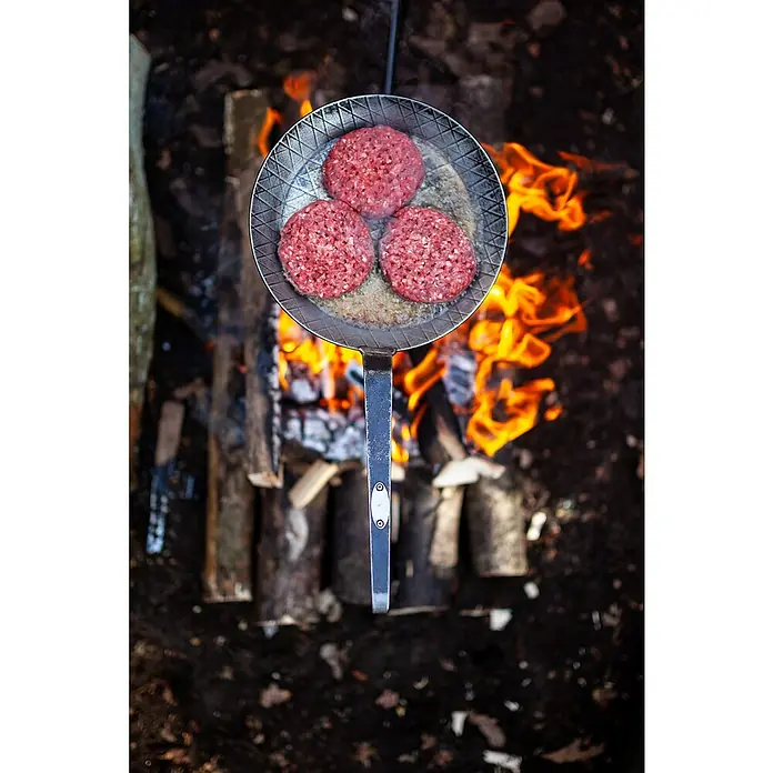 Petromax Grillpfanne - Durchmesser: 24,5 cm - 32 cm, Höhe: 17 cm, Eisen - Drei rohe Burger-Patties brutzeln in einer Pfanne über einem Lagerfeuer, bereit, gegrillt und serviert zu werden.
