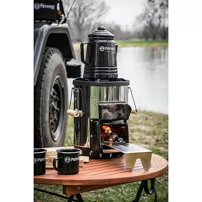 Petromax Perkolator Perkomax - Schwarz, 1,3 l - Schwarze Petromax Perkolator auf brennender Petromax Atago Feuerschale, mit Tassen auf Holztisch, neben Fahrzeug und Wasser.