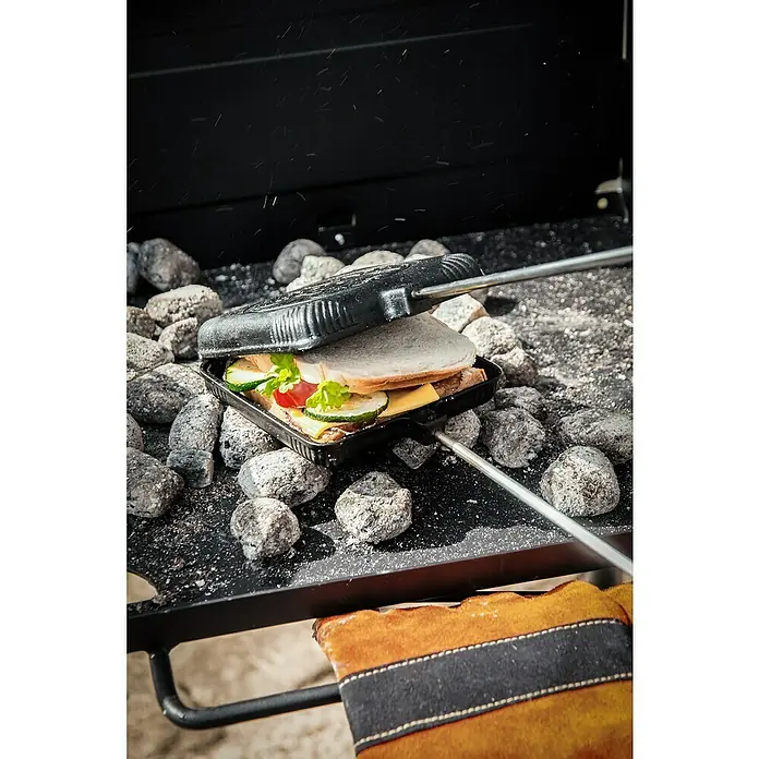 Petromax Sandwicheisen - L x B: 72,5 x 13,5 cm, Gusseisen - Sandwich-Eisen über Holzkohle. Das Sandwich-Eisen ist mit einem köstlichen Sandwich gefüllt, das auf dem Grill zubereitet wird.