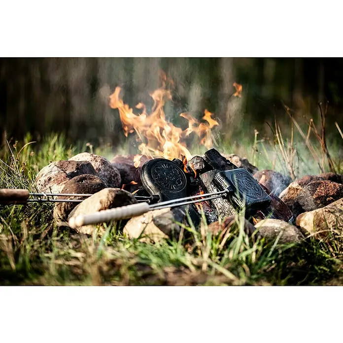 Petromax Sandwicheisen - L x B: 72,5 x 13,5 cm, Gusseisen - Das Bild zeigt zwei Stockbrot-Eisen über einem Lagerfeuer, umgeben von Steinen und Gras. Die Eisen sind ein rustikales Kochutensil für den Außenbereich.