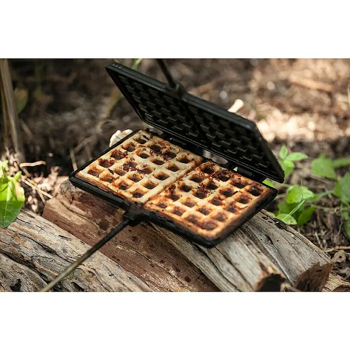 Petromax Waffeleisen - Länge: 72 cm, Gusseisen - Ein offenes Waffeleisen mit frisch gebackenen Waffeln, das auf einem rustikalen Holzscheit liegt. Perfekt für ein Picknick im Freien.