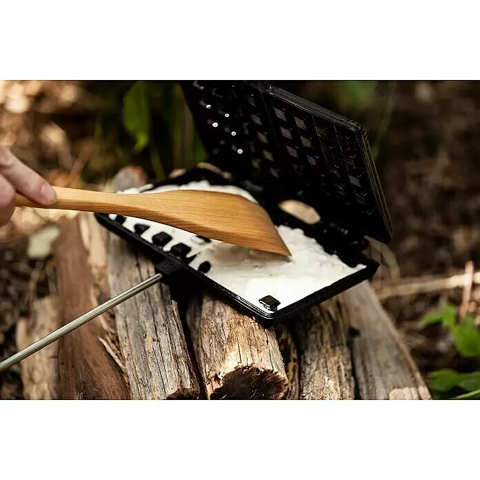 Petromax Waffeleisen - Länge: 72 cm, Gusseisen - Offenes dunkles Waffeleisen mit weißem Teig und Holzspatel, draußen verwendet. Eine Hand verteilt Teig in der Eisenform.