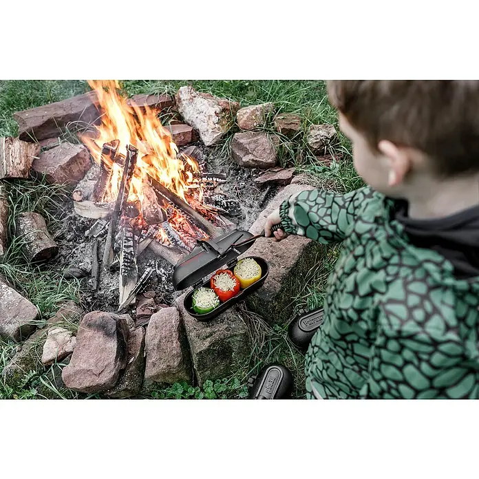 Petromax Kartoffelbräter - L x B x H: 29,1 x 17 x 15,4 cm, Gusseisen - Ein Kind bereitet gefüllte Paprika in einer gusseisernen Pfanne am Lagerfeuer zu. Das Essen wird über offenem Feuer gekocht.
