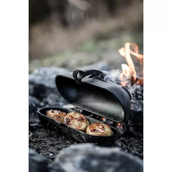 Petromax Kartoffelbräter - L x B x H: 29,1 x 17 x 15,4 cm, Gusseisen - Schwarzes Gusseisen-Sandwicheisen gart goldbraune Speisen über Lagerfeuer mit Rauch und Flammen.