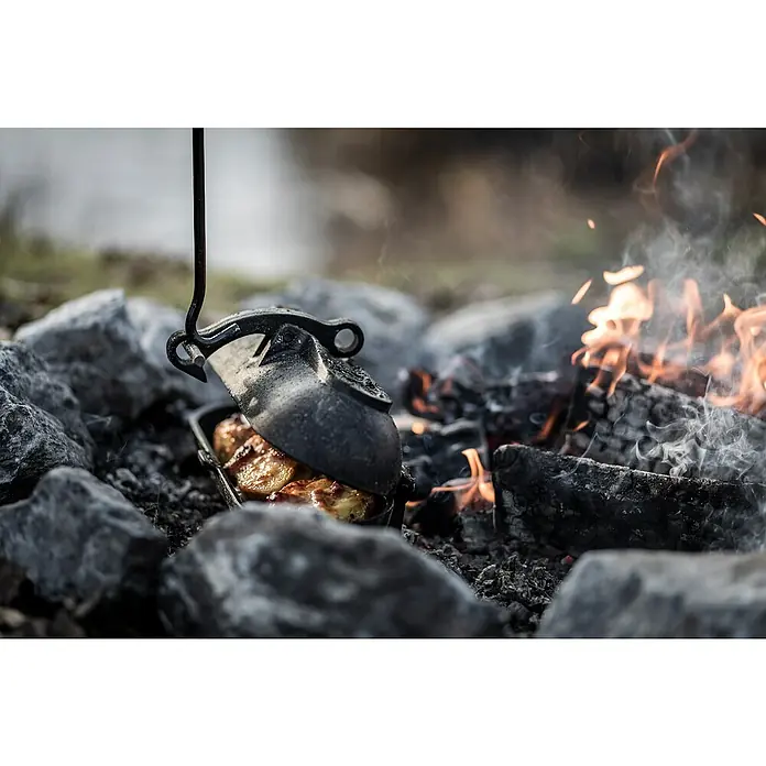 Petromax Kartoffelbräter - L x B x H: 29,1 x 17 x 15,4 cm, Gusseisen - Ein rustikales Lagerfeuer mit einem Dutch Oven, der an einem Haken hängt und köstliche Speisen über glühenden Kohlen zubereitet.