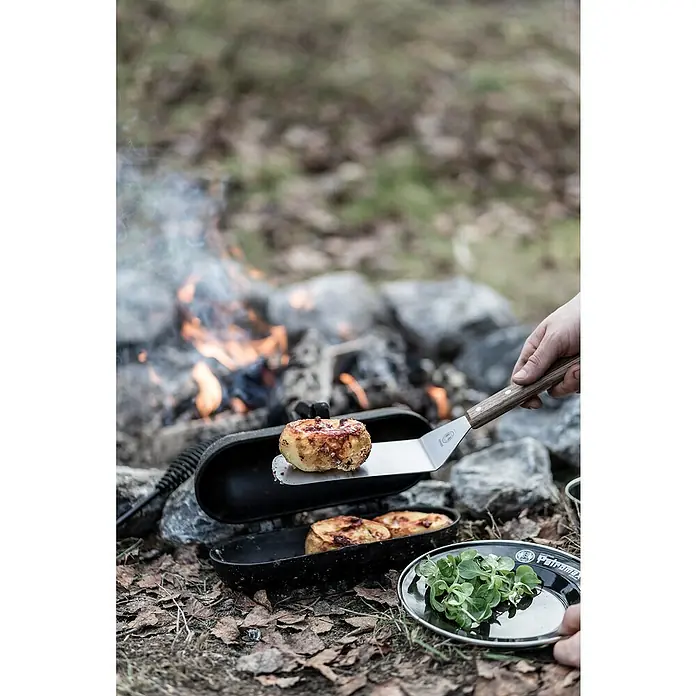 Petromax Kartoffelbräter - L x B x H: 29,1 x 17 x 15,4 cm, Gusseisen - Ein herzhaftes Gericht wird über offenem Feuer zubereitet, mit einem Pfannenwender und einem Teller mit frischen Kräutern.