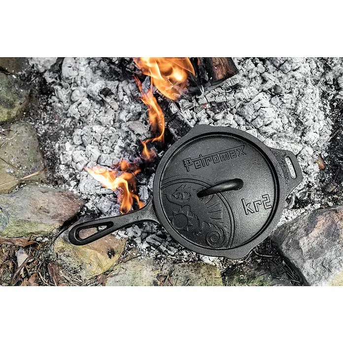 Petromax Kasserolle - Gusseisen, Durchmesser: 22 cm - Petromax Feuerpfanne mit Deckel, Modell kR2, neben einem Lagerfeuer. Ideal für Kochen im Freien, robust und vielseitig einsetzbar.
