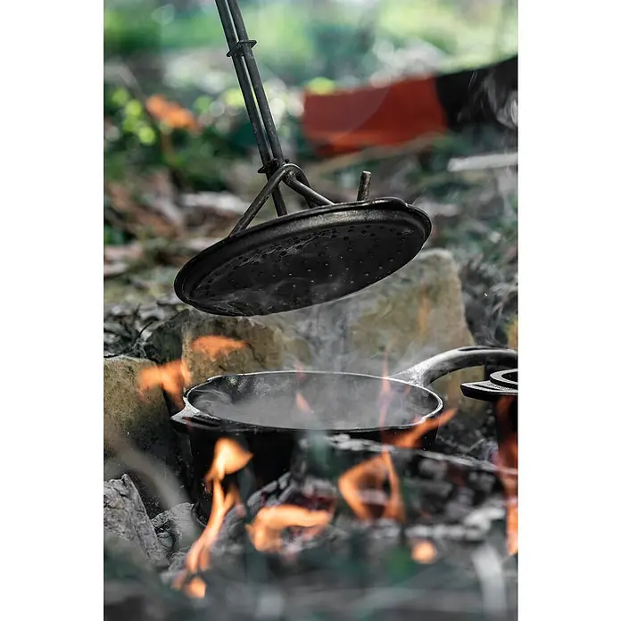 Petromax Kasserolle - Gusseisen, Durchmesser: 22 cm - Kochen im Freien: Ein schwarzer Topf steht über einem Lagerfeuer, während ein Deckel mit Löchern daneben hängt.