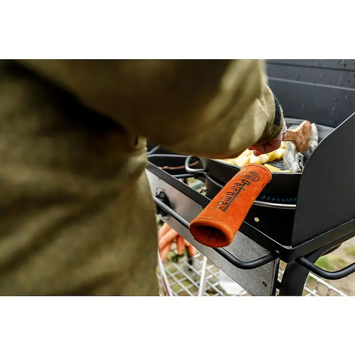 Petromax Handschutzgriff - Passend für: Petromax Grillpfannen, Leder/Aramidfasern - Detailaufnahme eines Kochs am Grill, der mit einem Griffschutz von Ferromax hantiert, während er Speisen zubereitet.
