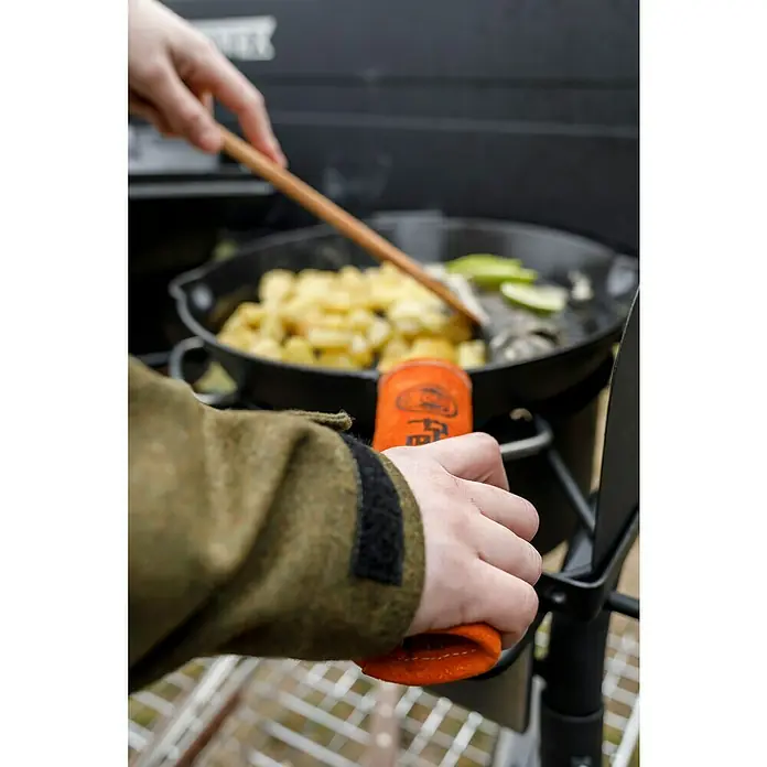Petromax Handschutzgriff - Passend für: Petromax Grillpfannen, Leder/Aramidfasern - Blick auf eine Bratpfanne mit Lebensmitteln auf einem Herd, eine Hand hält den Griff mit einem Topflappen, während die andere mit einem Holzlöffel umrührt.