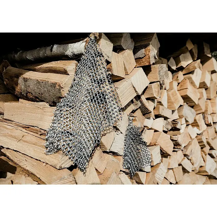 Petromax Grillreiniger Ringreiniger - 20 x 20 cm, Edelstahl - Nahaufnahme von zwei Kettenhemden, die an einem Stapel Holzscheite gelehnt sind, wobei die Metallstruktur und das Holz hervorgehoben werden.