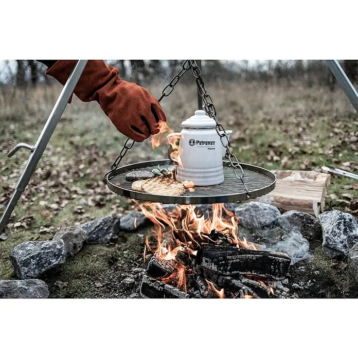 Petromax Grillrost für Dreibeinständer - Durchmesser: 50 cm, Stahl - Petromax Grillrost mit Feuerstelle, an dem Speisen zubereitet werden, und eine Hand mit einem Ofenhandschuh, die ein Gericht bewegt.