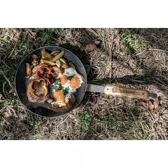 Petromax Haltegriff - L x B x H: 16 x 3,9 x 1,4 cm, Holz - Metallpfanne mit Holzgriff und Gravur, gefüllt mit gekochtem Essen, auf trockenem Gras liegend.