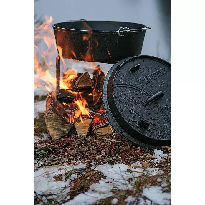 Petromax Dutch Oven - Ø x H: 36,4 x 16 cm, Gusseisen, 5,5 l, Planer Boden - Schwarzer gusseiserner Dutch Oven auf einem Gestell über einem Lagerfeuer, der Deckel ruht auf dem verschneiten Boden. Der Topf heizt, der Deckel zeigt dekorative Muster.