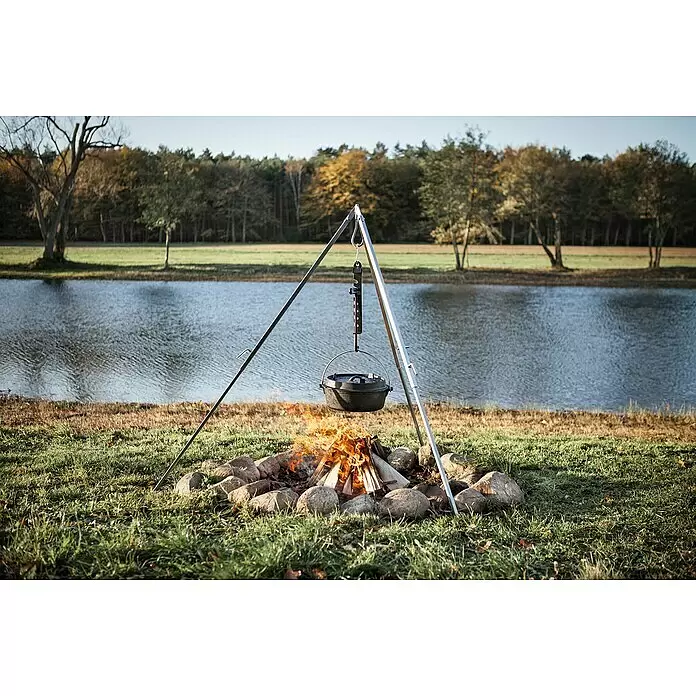 Petromax Dutch Oven - Ø x H: 36,4 x 16 cm, Gusseisen, 5,5 l, Planer Boden - Schwarzer gusseiserner Dutch Oven hängt an Metall-Dreibein über Lagerfeuer, kocht im Freien an See mit Herbstbäumen.