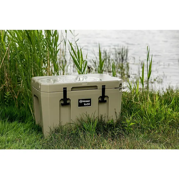 Petromax Kühlbox - Sand, 50 l, L x B x H: 68,5 x 48,5 x 50 cm - Eine robuste Petromax kx50 Kühlbox in Khaki steht im Gras am Ufer, ideal für Outdoor-Abenteuer und zum Frischhalten von Lebensmitteln.