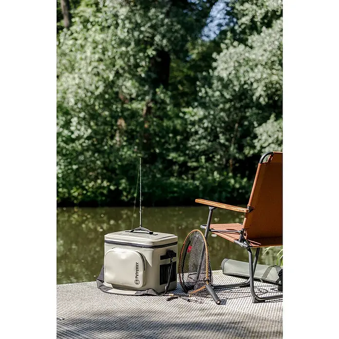 Petromax Kühltasche - Sand, 22 l, L x B x H: 42,5 x 34 x 35,5 cm - Ein Petromax Kühlbox mit einer Angelrute darauf steht neben einem Stuhl und einem Netz auf einer Plattform im Freien mit Bäumen im Hintergrund.