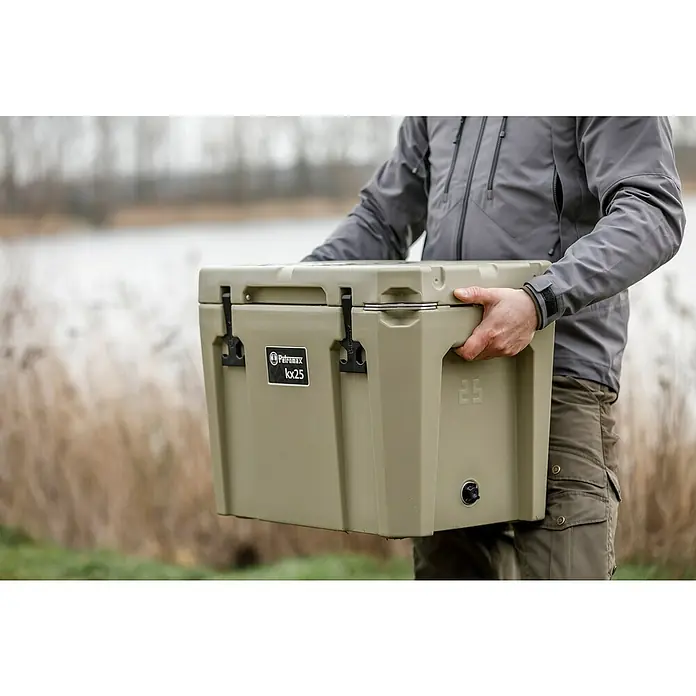 Petromax Kühlbox - Sand, 25 l, L x B x H: 58 x 40,5 x 45 cm - Eine Person trägt eine Petromax kx25 Kühlbox in Khaki vor einer unscharfen Naturkulisse. Die Kühlbox ist robust und ideal für Outdoor-Abenteuer.