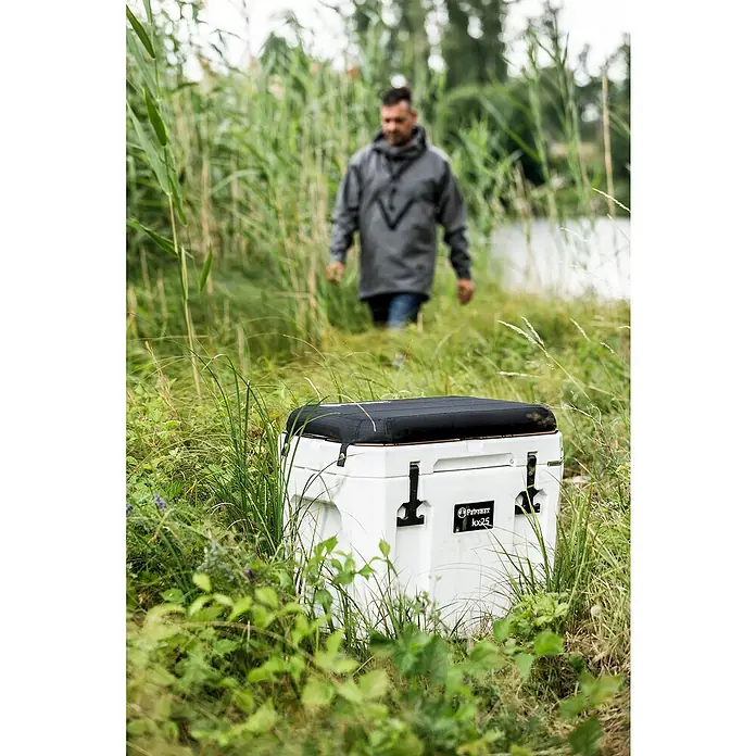 Petromax Sitzkissen - Passend für: Petromax Kühlbox 25 l, L x B x H: 38,5 x 56 x 5,5 cm, Schwarz - Der Petromax Kühlbox kx25 in Weiß steht im Gras, während eine Person im Hintergrund vorbeigeht. Perfekt für Outdoor-Abenteuer.