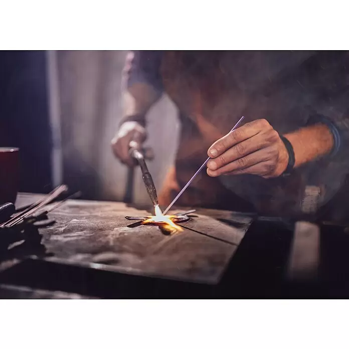 CFH Hartlötstab SH320 Silber - Schmelztemperatur: 670 °C - Nahaufnahme von Händen, die ein Metallobjekt mit einem Brenner und einem lila Schweißstab löten, wodurch ein helles orangefarbenes Glühen entsteht.