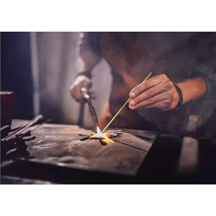 CFH Hartlötstab Neusilber - Ø x L: 2 x 333 mm, 5 Stk. - Detailaufnahme von einem Handwerker, der mit einem Schweißbrenner ein Metallobjekt bearbeitet, wobei die Hitze und die Präzision des Prozesses hervorgehoben werden.