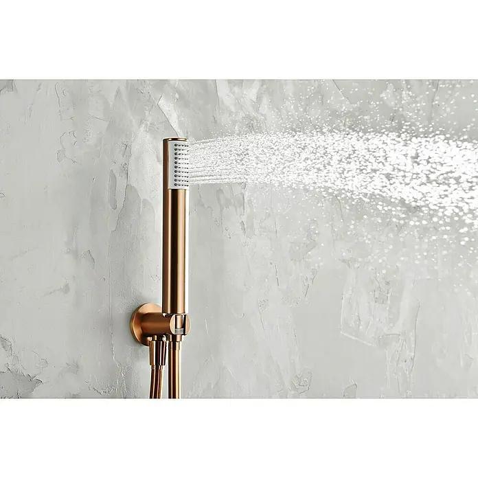 Camargue Natur Sistema de ducha empotrado - Con grifo monomando, Oro Rosa - Primer plano de un cabezal de ducha de latón rociando agua, montado en una pared gris texturizada. Diseño de baño elegante con un toque funcional.
