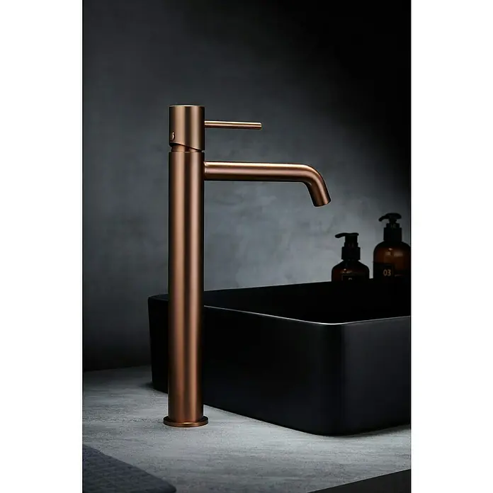 Camargue Natur Grifo de lavabo alto - Oro rosa - Elegante grifo de cobre junto a un lavabo negro. Diseño minimalista, perfecto para baños modernos, con elegantes dispensadores de jabón al fondo.