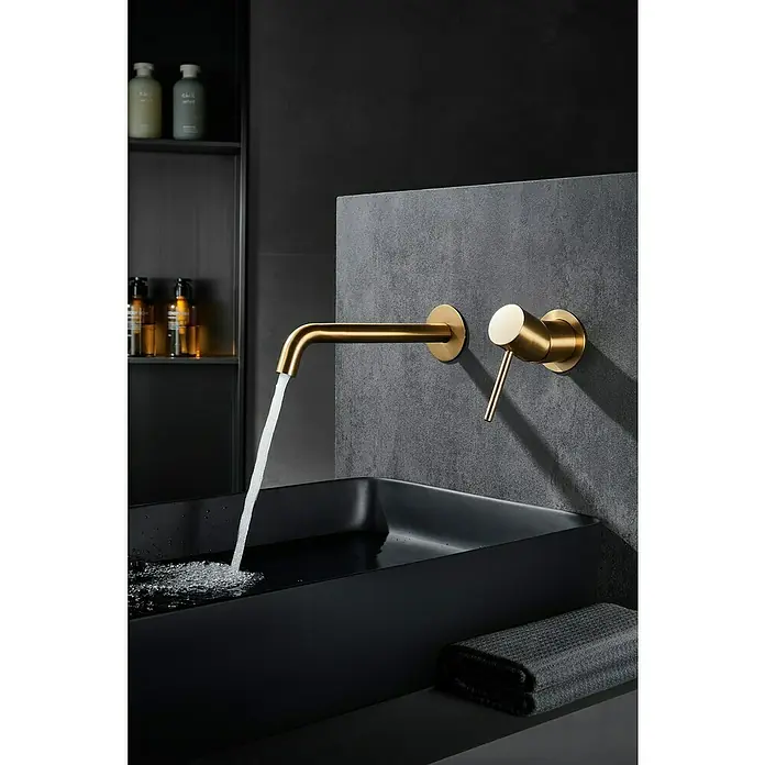 Camargue Natur Grifo de lavabo empotrado - Oro - Elegante grifo de baño dorado con diseño moderno. El grifo está montado en una pared oscura y alimenta agua a un lavabo negro.