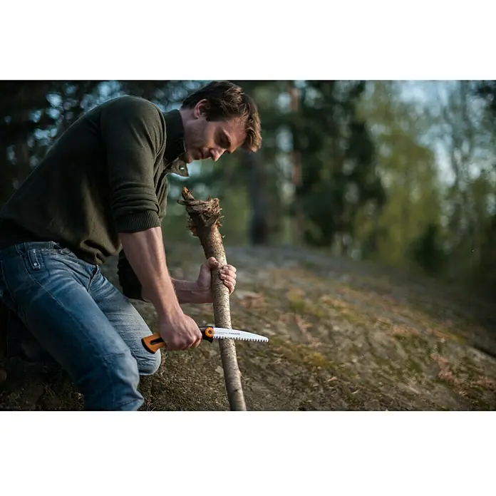 Fiskars Hand-Astsäge Xtract SW73 - Länge: 22,5 cm, Max. Aststärke: 12 mm, Stahl, Orange/Schwarz - Ein Mann benutzt eine Fiskars Handsäge, um einen Ast zu schneiden, während er in einer Waldumgebung kniet. Praktisch für Outdoor-Abenteuer.