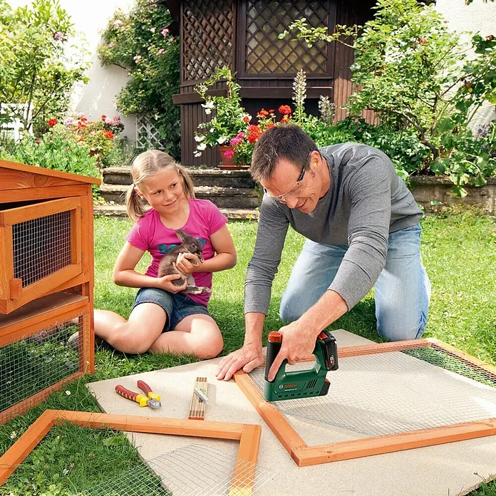 Bosch Power for All 18V Akku-Tacker UniversalTacker 18V-14 - 18 V, Ohne Akku, Klammertiefe: 6 mm - 14 mm - Mann, Mädchen und Kaninchenstallbau im Garten. Ein grüner Bosch Tacker ist im Einsatz, um Draht an einem Holzrahmen zu befestigen.