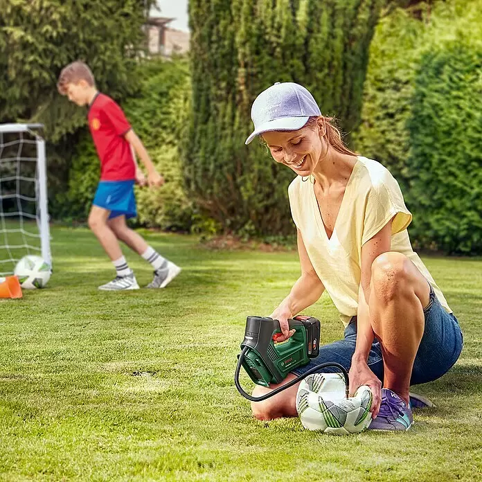 Bosch Power for All 18V Bomba de aire de batería UniversalPump - 18 V, 1 batería, 2 Ah, 10,3 bar - Una mujer usa un inflador portátil Bosch verde y negro para inflar un balón de fútbol blanco. Un niño juega al fondo.