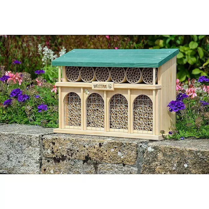 Dobar Insektenhotel Dublin - 44 x 13,5 x 30 cm, Holz, Braun/Grün - Ein hölzernes Bienenhotel mit grünem Dach, gefüllt mit Röhren und Gitter, auf einer Steinmauer in bunten Gartenblumen.