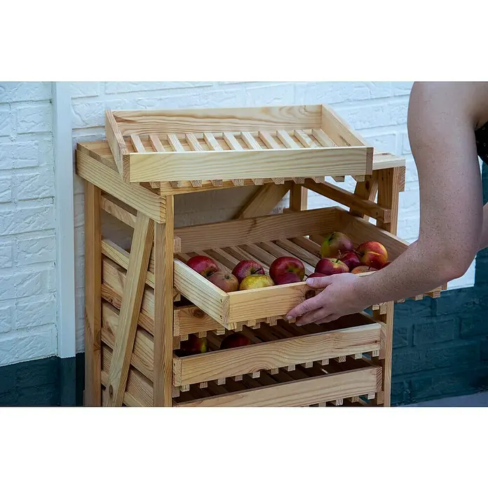 Dobar Kartoffel- & Obsthorde - L x B x H: 51 x 40,5 x 70,5 cm - Ein hölzernes Obstregal mit Äpfeln, wobei eine Person eine Schublade herauszieht. Das Regal steht vor einer weißen Backsteinmauer.