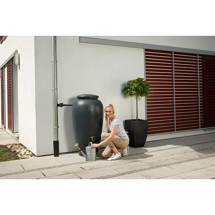 4rain Regenspeicher Amphore - 220 l, Graphite grey - Eine Frau befüllt eine Gießkanne an einem grauen Regenspeicher vor einem Haus mit braunen Fensterläden.