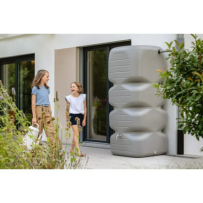 Garantia Basic Line Regentonne Cubus - 1.000 l, Betongrau - Zwei Mädchen stehen neben einem grauen Regenspeicher, der an der Hauswand befestigt ist. Die Regentonne hat einen Wasserhahn und einen Zulauf.