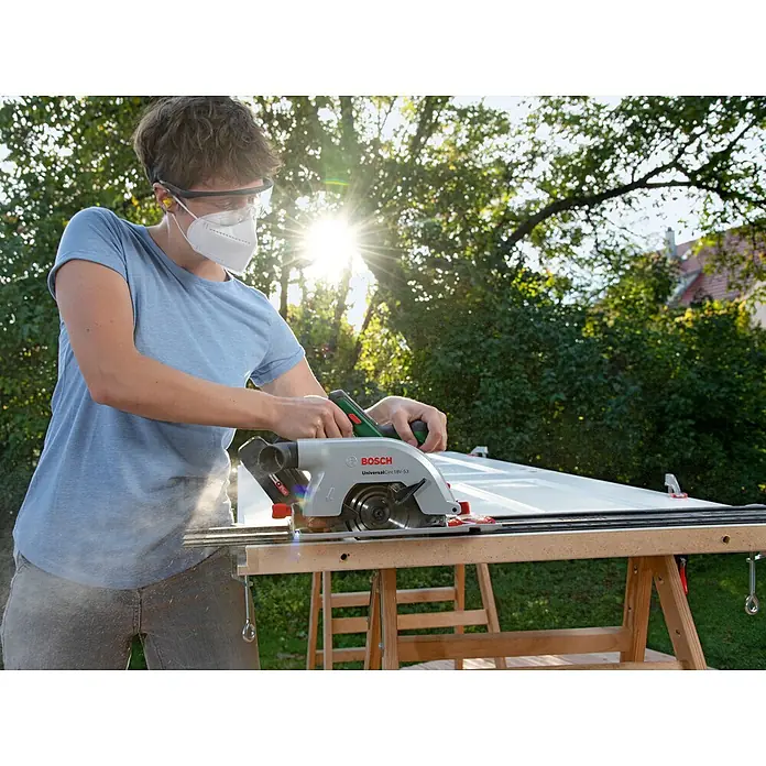 Bosch Power for All 18V Akku-Handkreissäge UniversalCirc 18V-53 - 18 V, Ohne Akku, 4.300 U/min, Durchmesser Sägeblatt: 160 mm - Eine Person benutzt eine Bosch Kreissäge, um ein Werkstück zu bearbeiten, wobei sie Schutzbrille und Maske trägt.