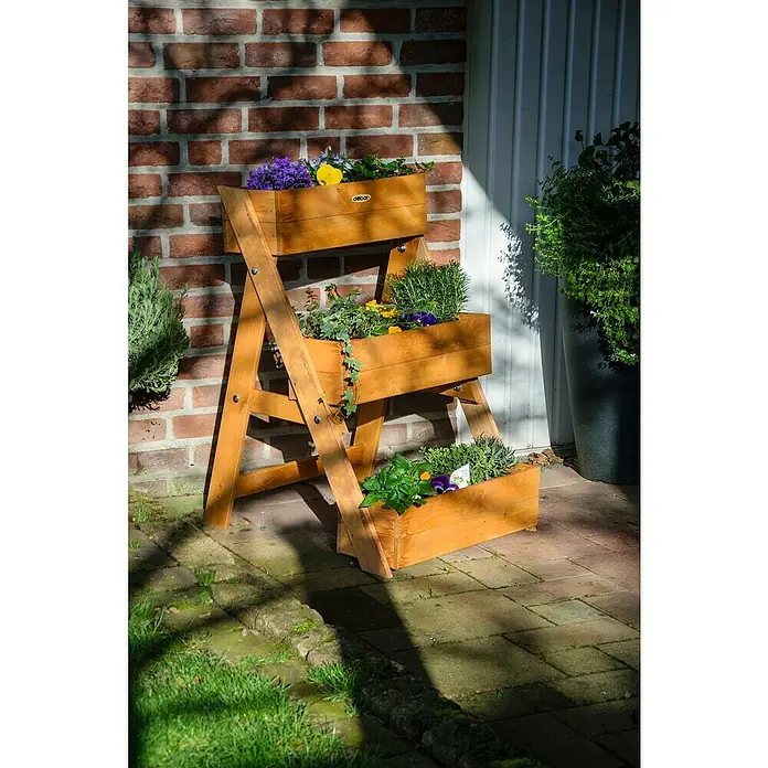 Dobar Pflanzkasten - L x B x H: 60 x 70 x 90 cm, Holz, Braun - Ein dreistufiger Holzpflanzenständer mit verschiedenen Blumen und Kräutern in Holzkisten, der vor einer Ziegelmauer und einer Tür steht.