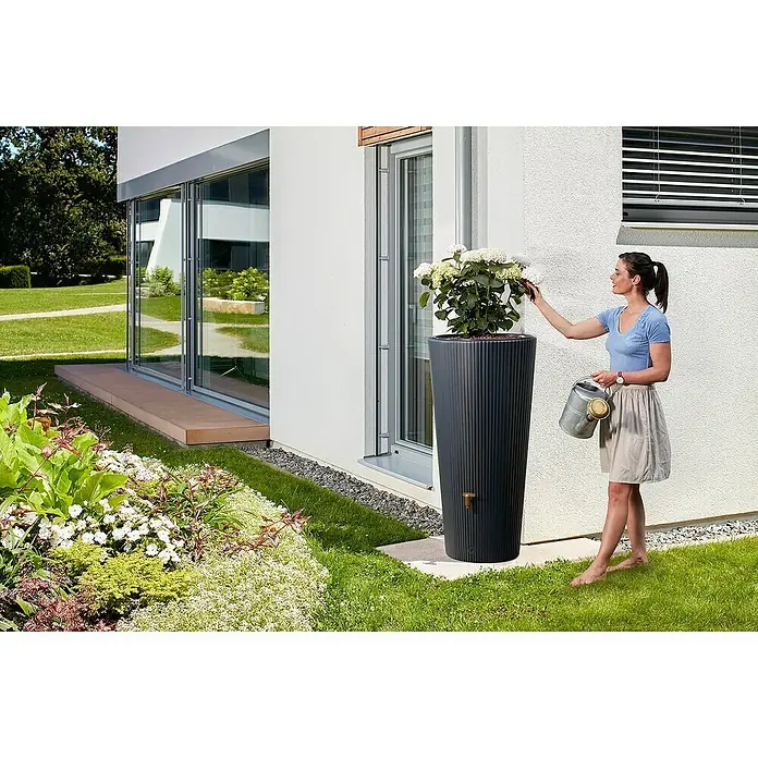 4rain Regenspeicher Vaso Decor 2in1 - 220 l, Graphite grey - Ein schlanker, gerippter Regenspeicher in Anthrazit mit einer Frau, die eine Pflanze im Garten gießt, fügt sich nahtlos in die moderne Gartengestaltung ein.