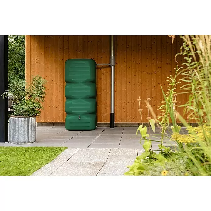 Garantia Basic Line Regentonne Cubus - 1.000 l, Dunkelgrün - Grüner Regentank mit Hahn, an ein Fallrohr angeschlossen, auf einer Terrasse an einer Holzwand zur Gartenbewässerung.