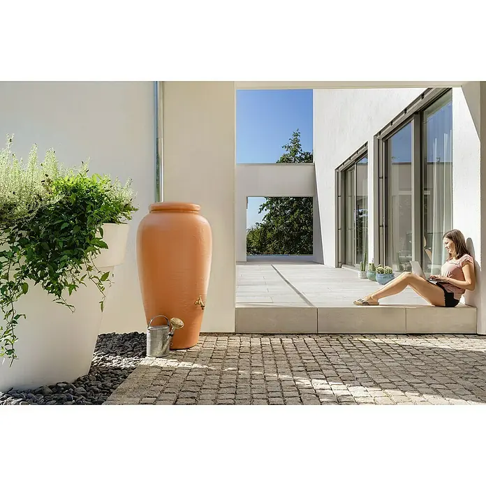 Garantia Classic Line Regenspeicher Amphore - 240 l, Terrakotta - Eine terracottafarbene Wassersäule steht neben einem Pflanzgefäß, während eine Frau auf einer Terrasse mit einem Laptop sitzt.