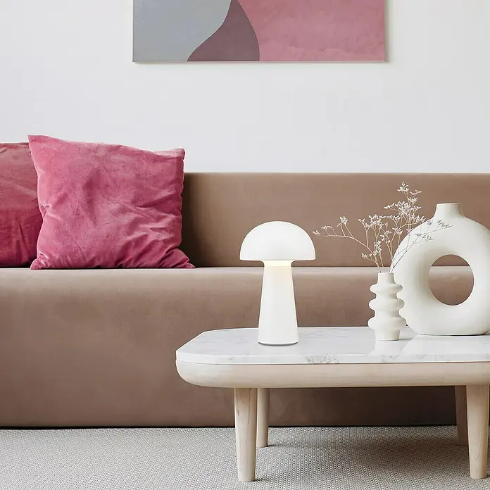 Trio Leuchten Akku-Tischleuchte Lennon - 2 W, Weiß, Warmweiß - Eine moderne Pilzlampe aus weißem Glas steht auf einem Tisch neben einer Couch und Vasen.