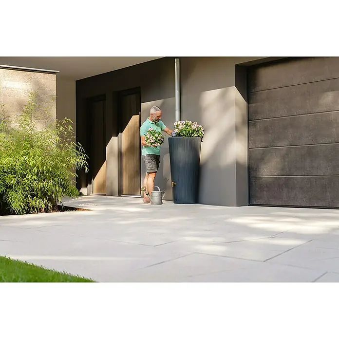 4rain Regenspeicher Vaso Decor 2in1 - 220 l, Graphite grey - Ein Mann bepflanzt eine schlanke, geriffelte Regentonnen-Säule mit rosa Blüten. Der Regenspeicher steht an einer Hauswand neben einer Regenrinne.