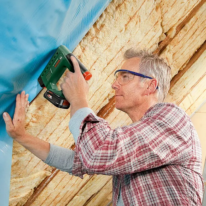 Bosch Power for All 18V Akku-Tacker UniversalTacker 18V-14 - 18 V, Ohne Akku, Klammertiefe: 6 mm - 14 mm - Ein Mann befestigt mit einem grünen Bosch Akku-Tacker blaue Folie an einer Dachschräge mit Dämmwolle.