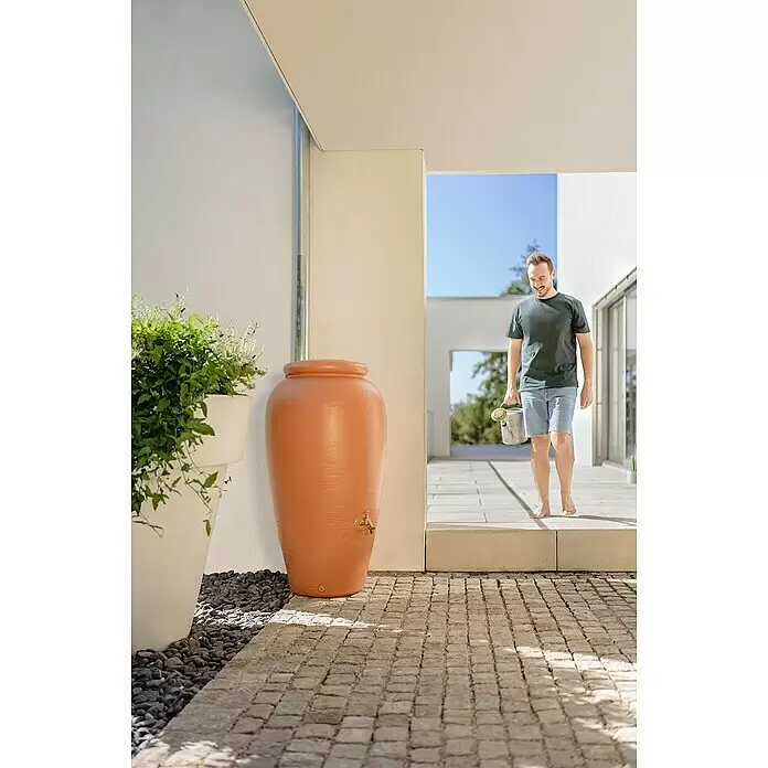 Garantia Classic Line Regenspeicher Amphore - 240 l, Terrakotta - Terrakotta-Regentonne sammelt Wasser vom Fallrohr; ein Mann mit Gießkanne kommt, um sie zur Gartenbewässerung zu nutzen.