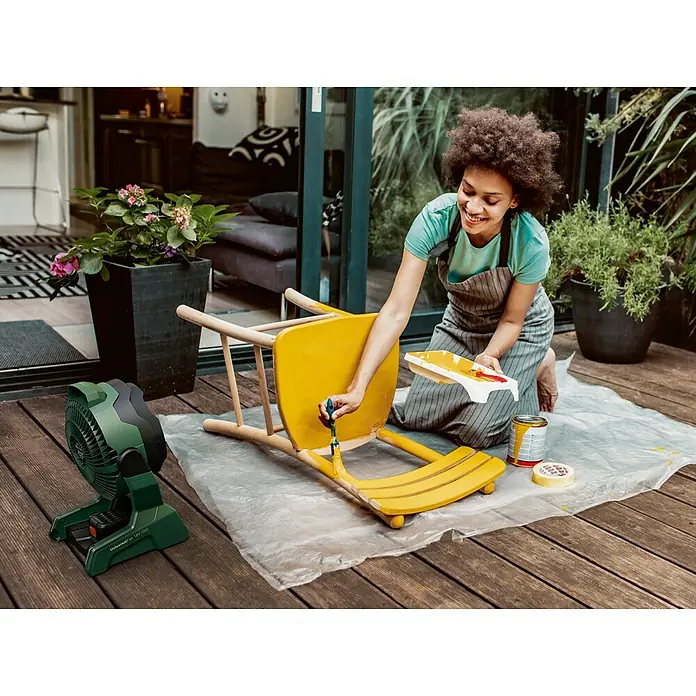 Bosch Power for All 18V Akku-Ventilator UniversalFan 18V-1000 - 18 V, 1 Akku, 2 Ah, Luftmenge: 1.000 m³/h - Eine Frau streicht einen Holzstuhl gelb mit einem Akku-Ventilator im Vordergrund, der für eine gute Luftzirkulation sorgt.