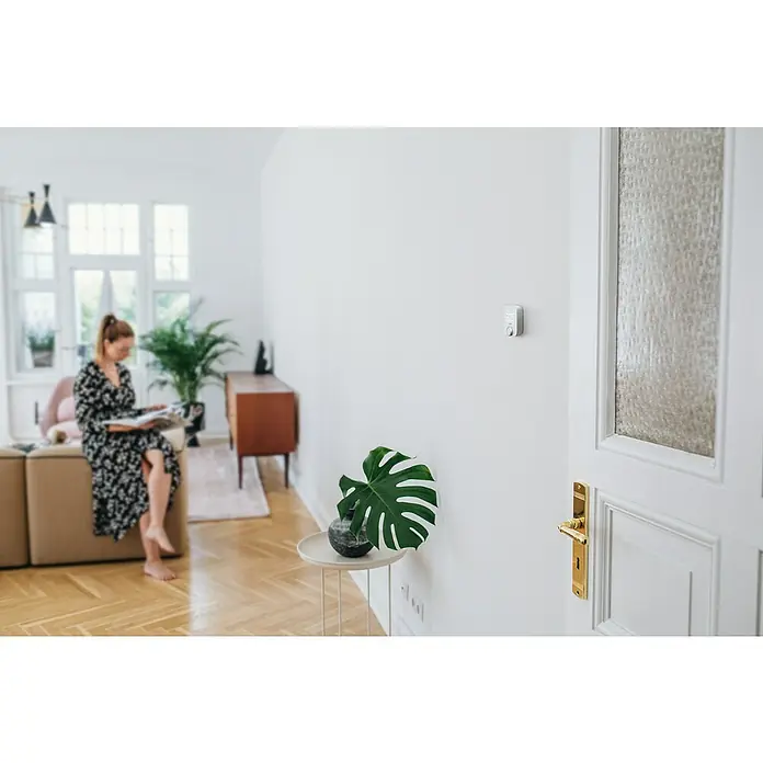 Bosch Smart Home Raumthermostat II Fußbodenheizung - 230 V, LED-Display, L x B x H: 35 x 86 x 87 mm - Ein Thermostat ist an einer weißen Wand in einem hellen Raum montiert, neben einer verzierten Tür und einer Zimmerpflanze.